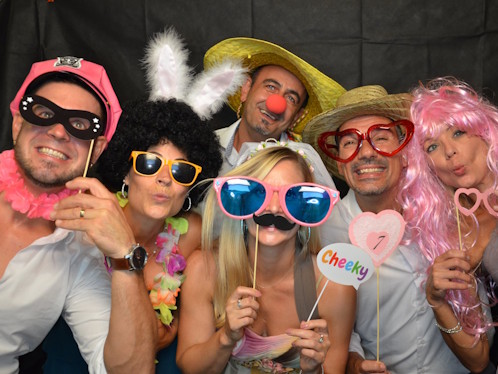 Gruppe von Erwachsenen mit bunten Verkleidungen und Foto‑Booth‑Accessoires, die gemeinsam ein lustiges Erinnerungsfoto aufnehmen