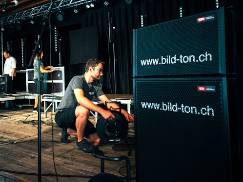 Techniker richten auf einer Bühne professionelle Eventtechnik ein, mit Lautsprechern, Kabelmanagement, Mikrofonen und Equipment-Cases während des Aufbaus für eine Veranstaltung.