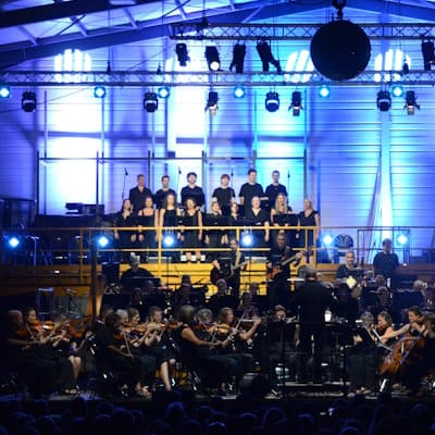 • Ein quadratischer Bildausschnitt des Konzerts „Epic Rock Symphony“ in der Werkhalle der Kurt Eberle AG in Roggwil-Arbon. Im Fokus steht der gemischte Chor in schwarzer Kleidung auf einer erhöhten Tribüne sowie die Rockband im Hintergrund. Davor sieht man den Dirigenten von hinten und einen Teil der Streichersektion des Sinfonischen Orchesters Arbon. Die Szene ist von oben intensiv blau und weiß beleuchtet, wobei eine große Spiegelkugel am oberen Rand sichtbar ist.